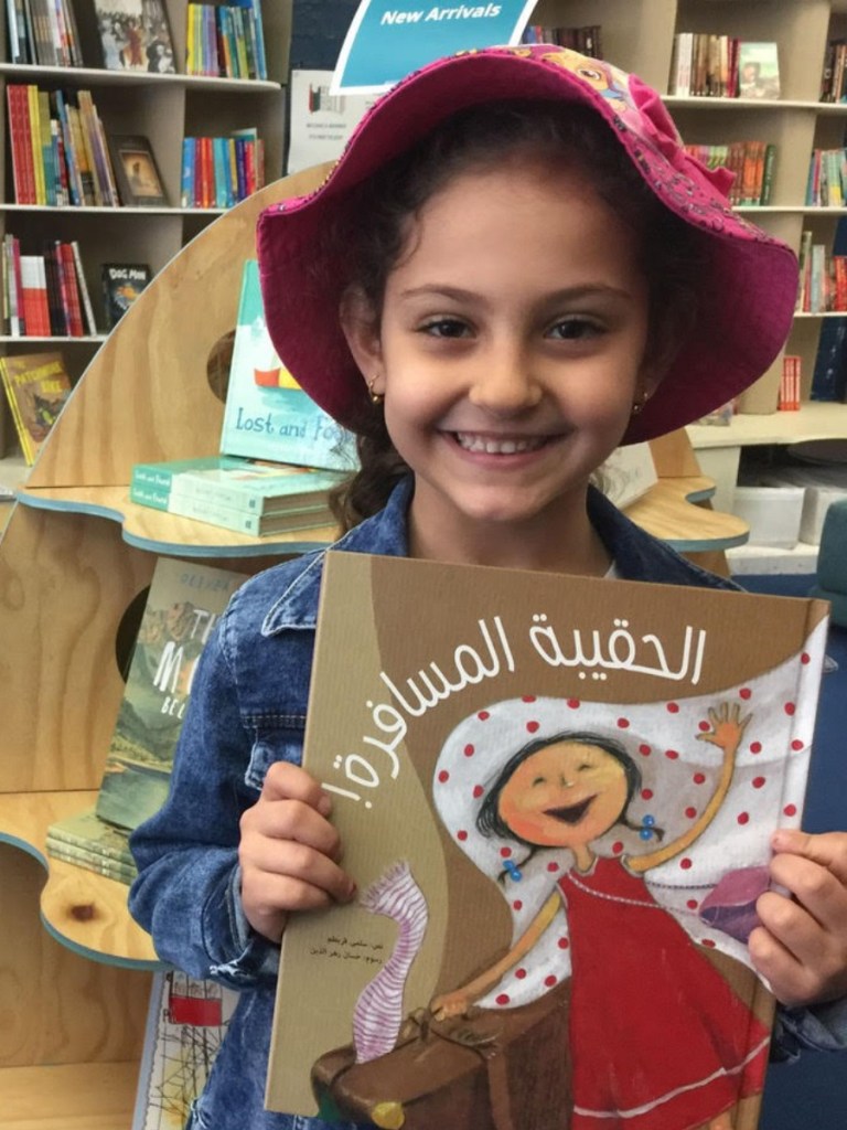 Girl in a pink hat and blue jacket holds a book with a character in a red dress.