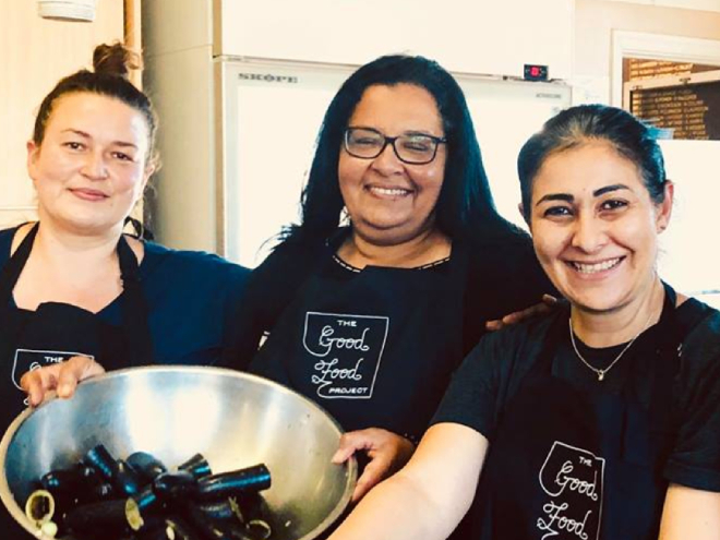 Three women in black shirts and apron stand in front of a bowl of food.