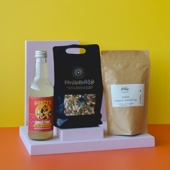 Yellow wall, orange floor, from L-R - a bottle of ginger tonic, a 250g pouch of Persian brekkie in black packaging with a clear window on the bottom, a 60g tea pouch