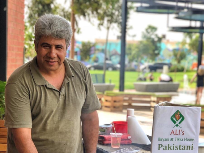 Man in a green shirt in front of a bbq with skewers on it, and a sign reading 'Pakistani food' on it nearby