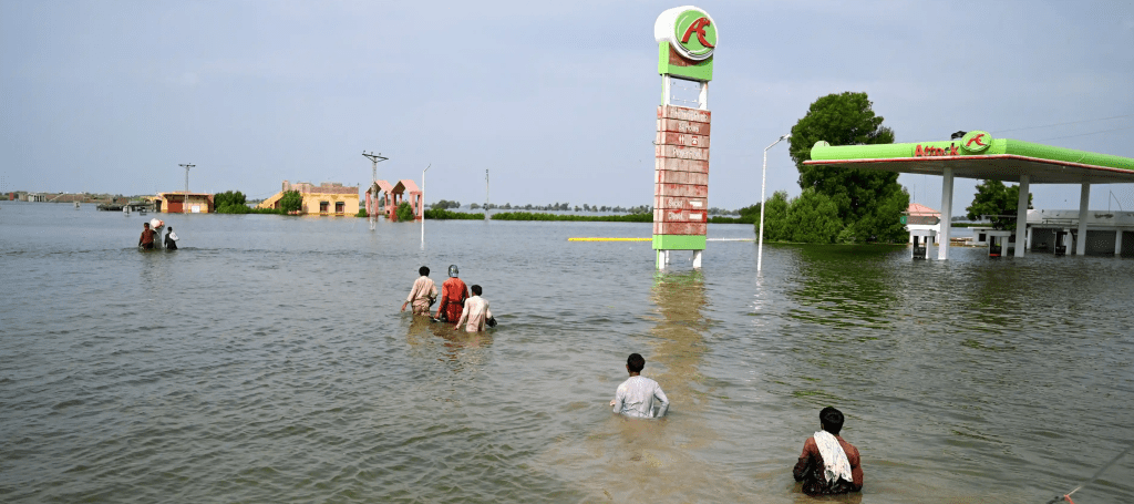 Pakistani people wading through flood waters that reaches their waists. A gas service station is half submerged nearby under water that spans out across the entire town, into the horizon.