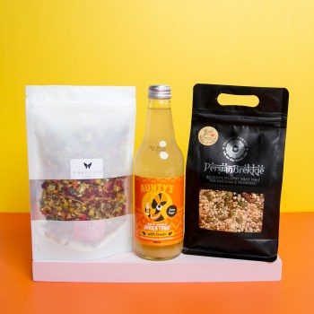 Yellow and orange backdrop. On a pink platform, there's a white tea pouch, a bottle of ginger tonic and a Persian brekkie pouch.