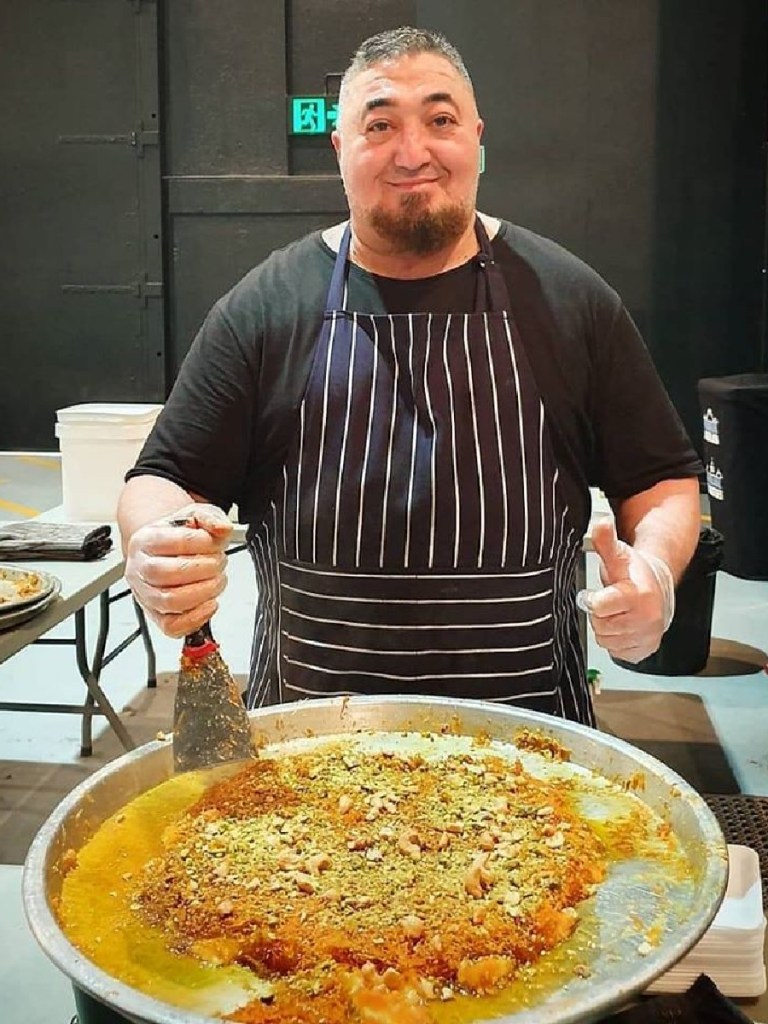 Man in a striped apron stands in front of fresh knafeh on a silver platter