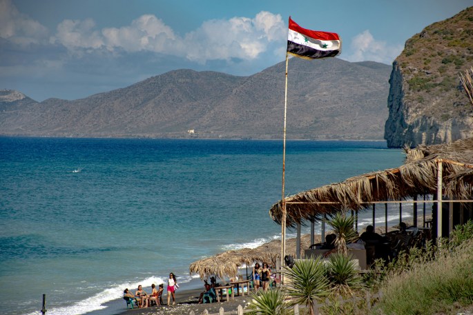 The Syrian flag waving atop a pole, with mountains & the sea in the background.